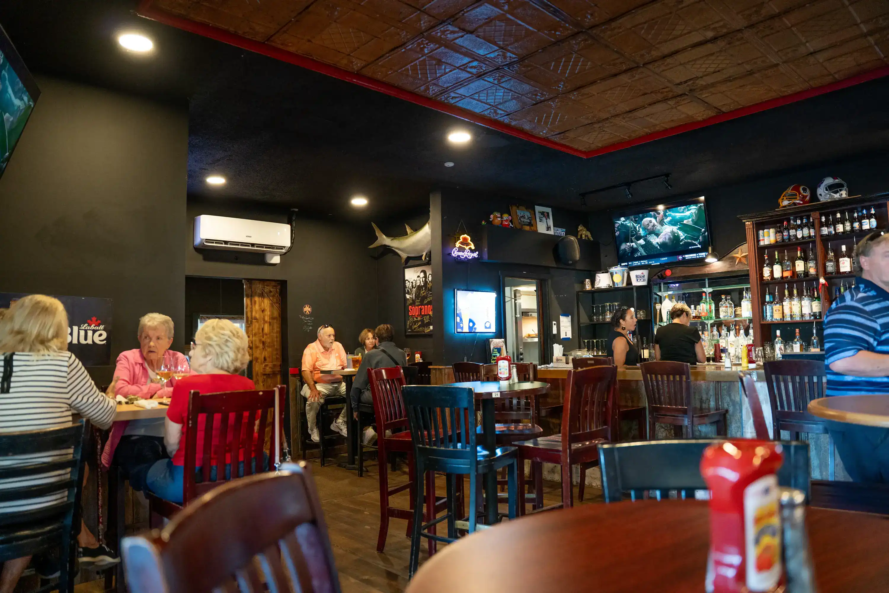 Sports bar dining area with dark walls, decorative tin ceiling, customers seated at tables, and full bar with mounted TVs