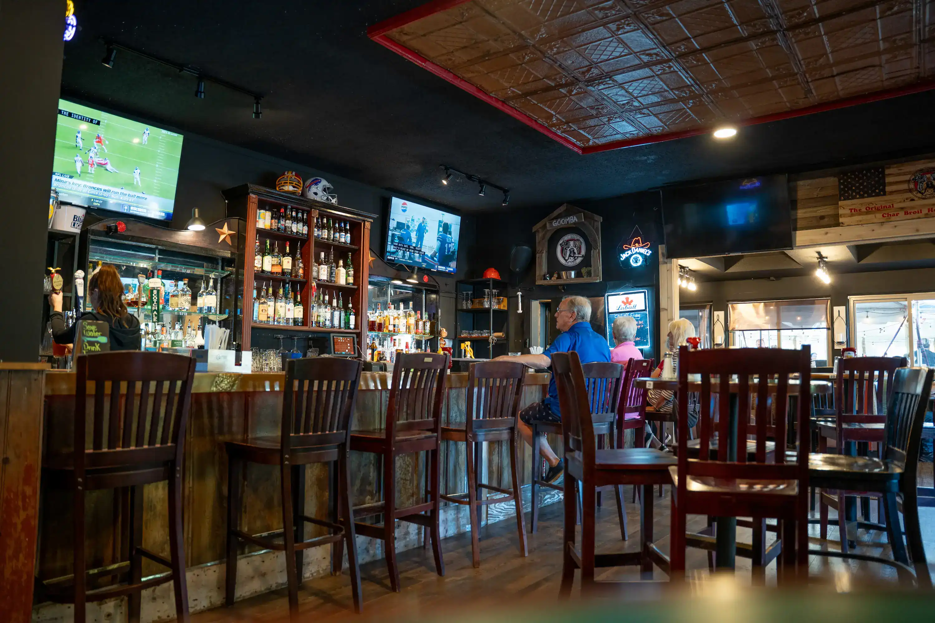 Bar interior with wooden stools, liquor shelves, multiple TV screens, and patrons seated at counter