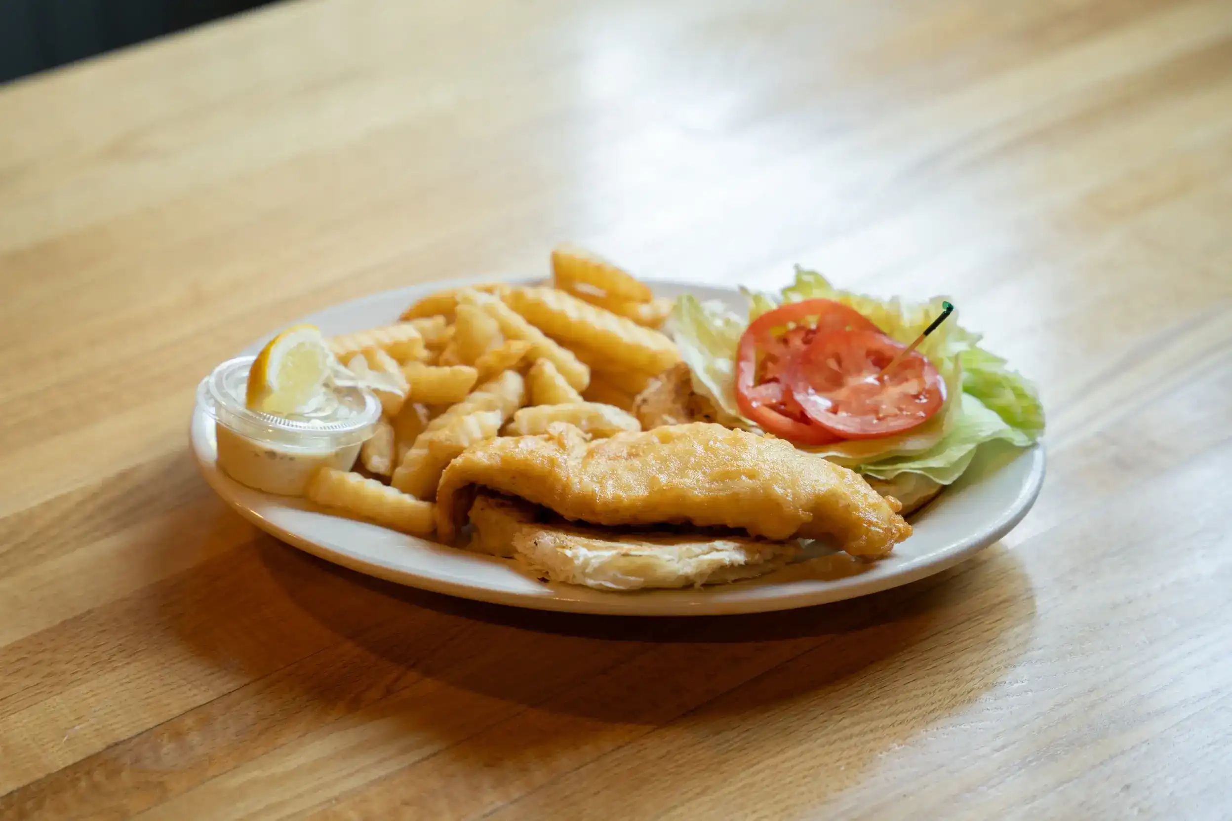 Battered fish fillet with french fries, lettuce, tomato, and lemon wedge on white plate