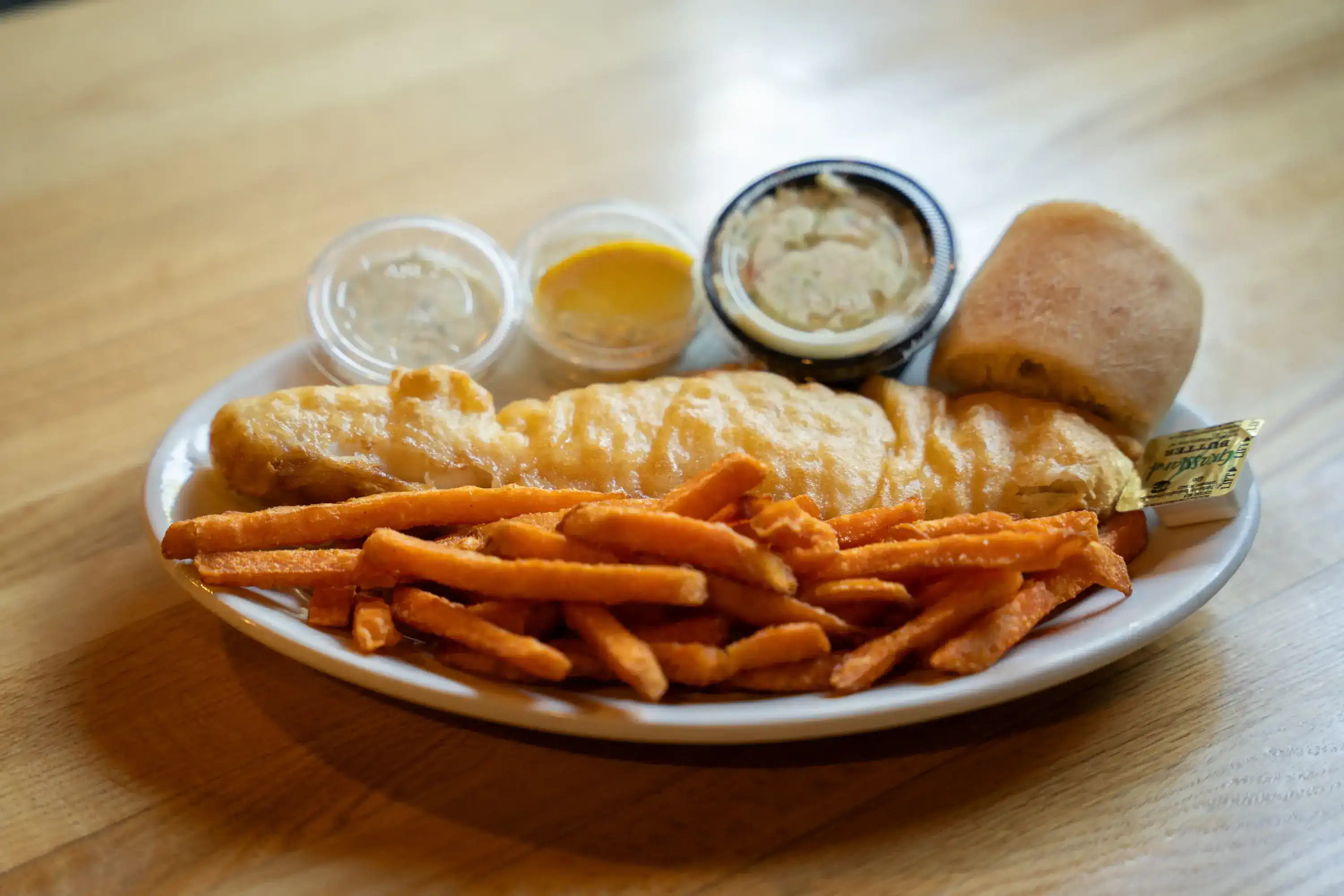 Fried fish fillets with sweet potato fries, dinner roll, and condiments on white plate