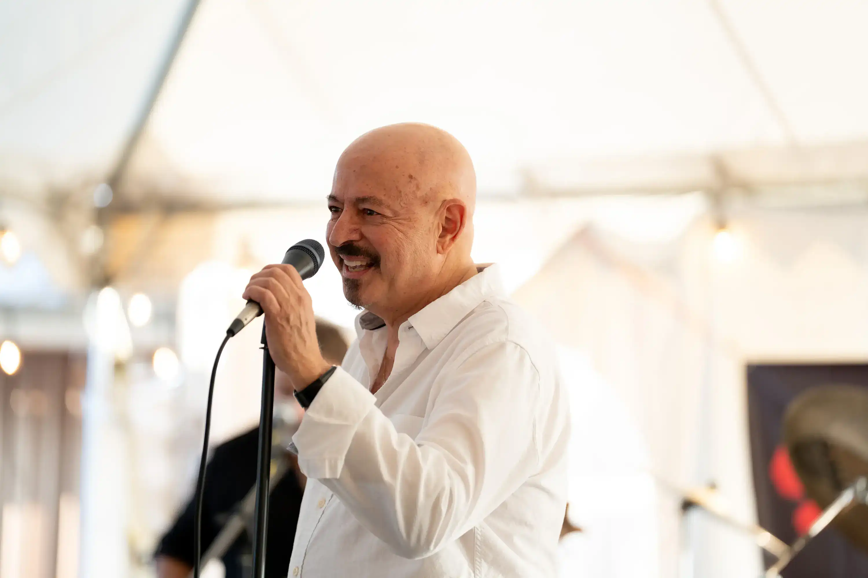Bald singer in white shirt performing with microphone under white tent at restaurant event