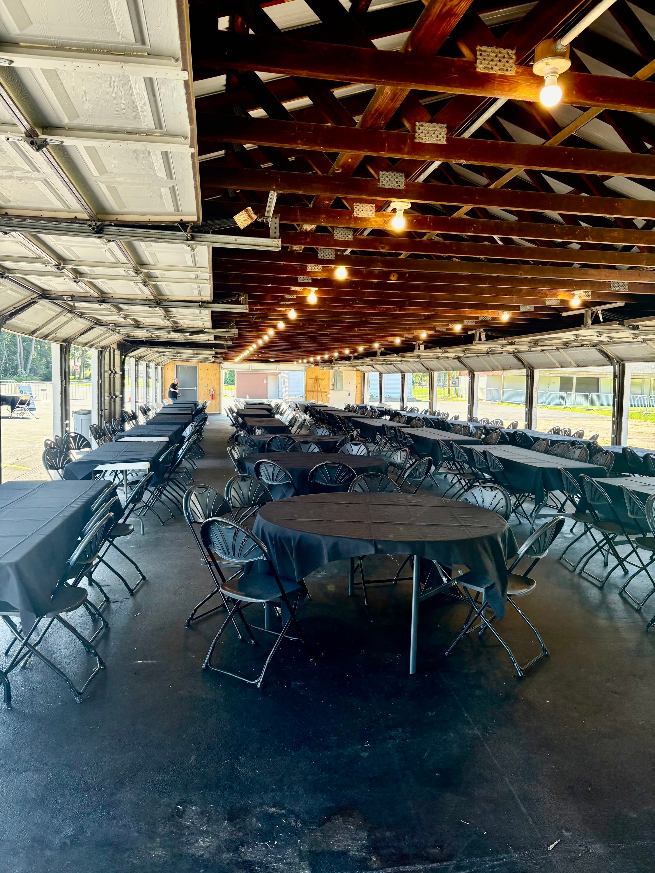 Open-air pavilion with round tables, black folding chairs, and exposed wooden beam ceiling with string lights