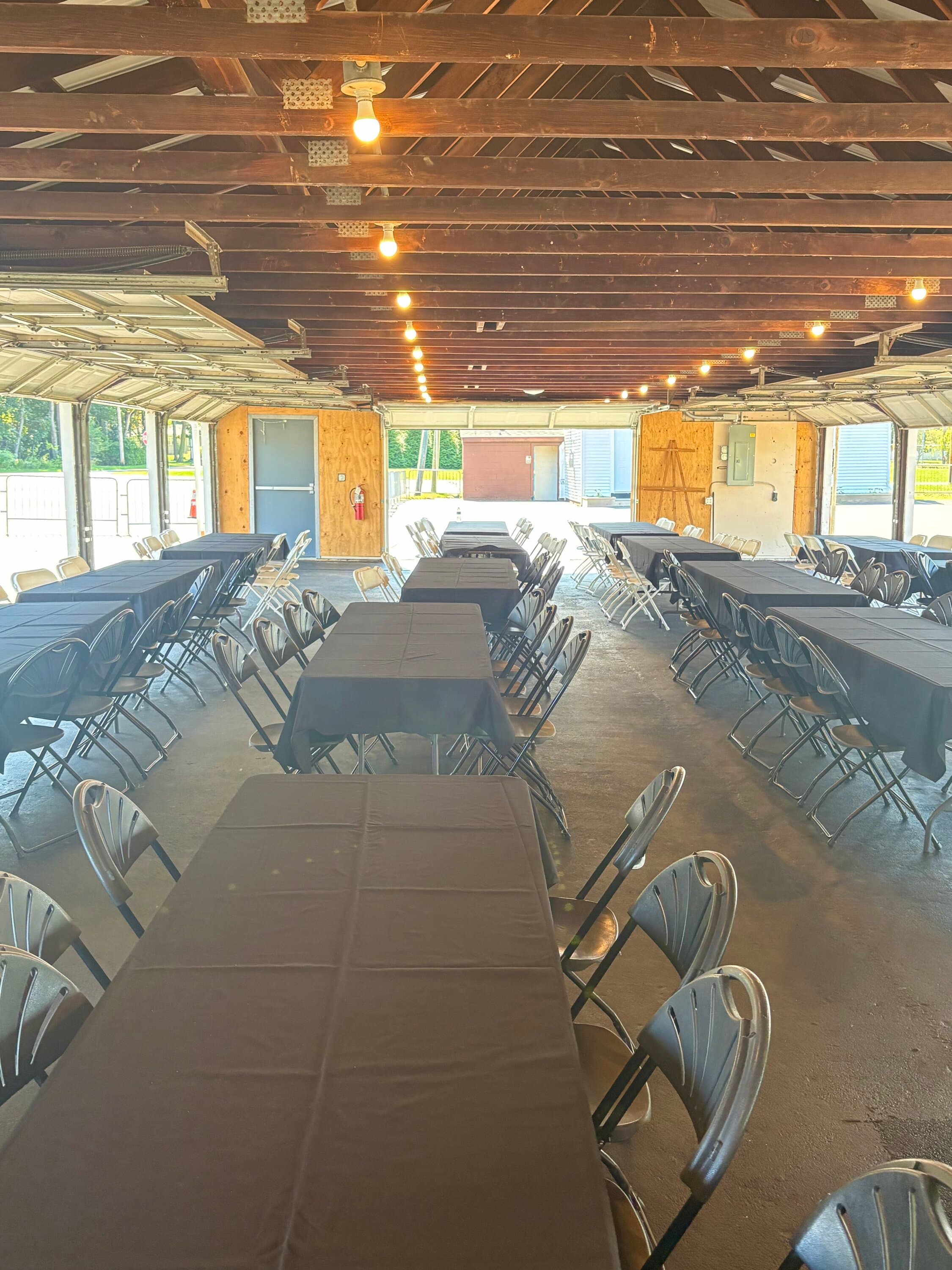 Open-air pavilion with rows of tables covered in dark linens and metal folding chairs, exposed beam ceiling with string lights