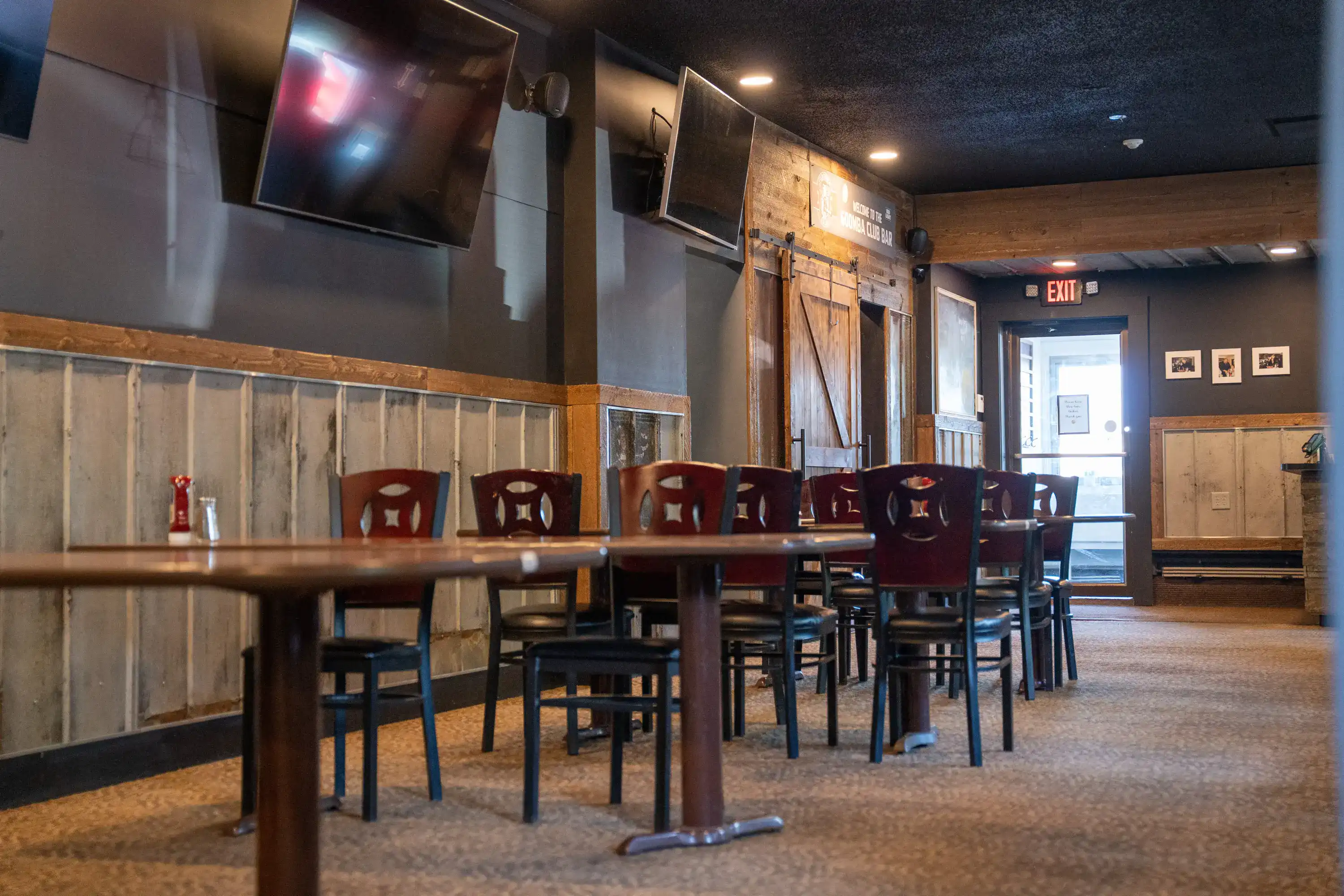 Restaurant dining room with wooden tables, dark chairs, mounted TVs, and rustic barn door decor