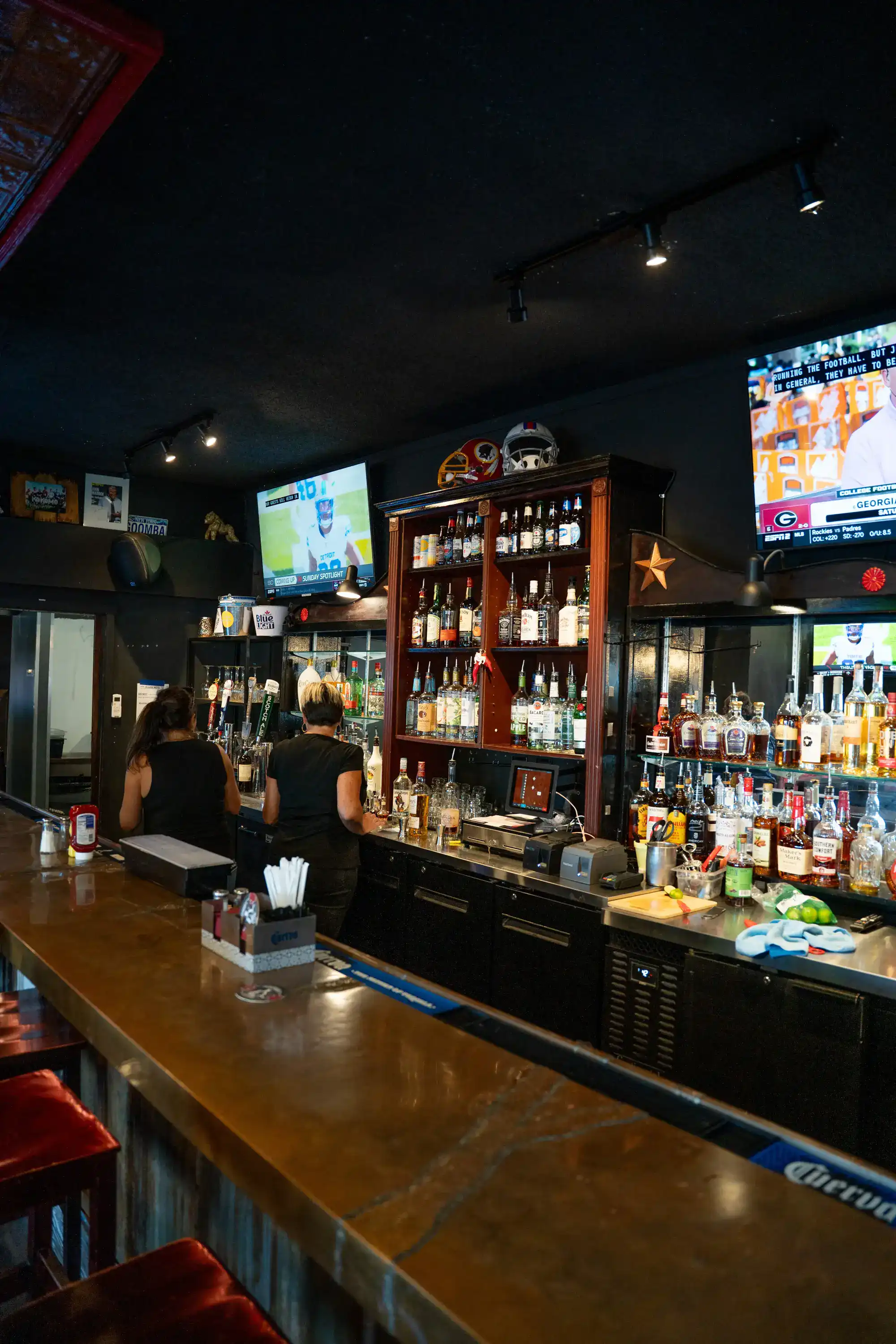 Bar area with sports memorabilia, multiple TV screens, and bartenders serving patrons