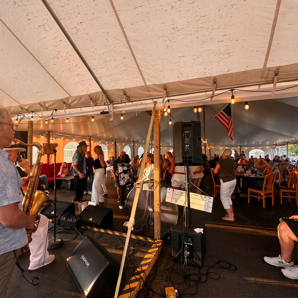 Crowded outdoor tent venue with a saxophonist performing, seated diners, string lights, speakers, and an American flag.