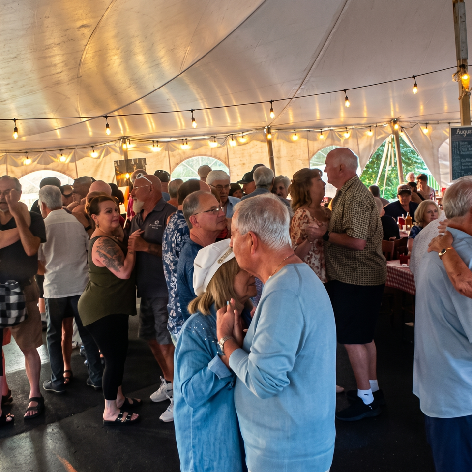 Guests dance beneath string lights inside a festive restaurant tent with picnic tables and a lively crowd.