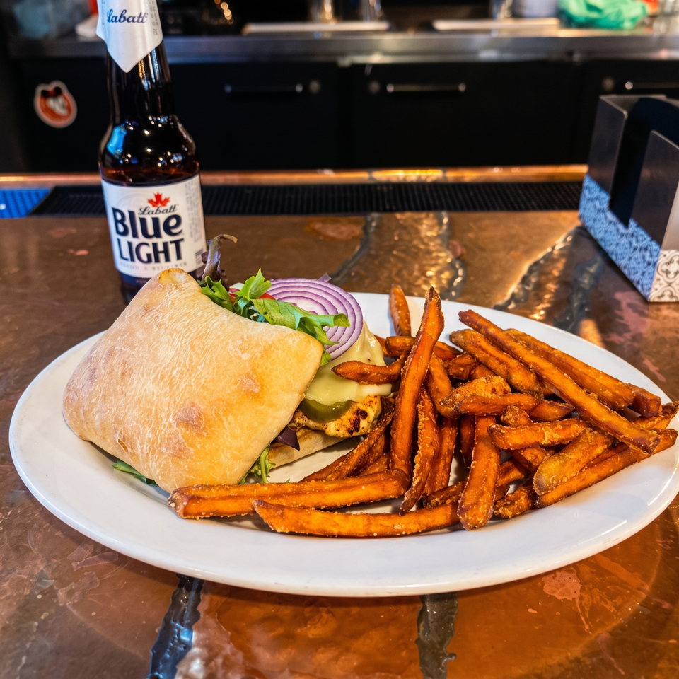 Grilled sandwich with lettuce, onions, pickles, and cheese served with sweet potato fries beside a Labatt Blue Light beer.